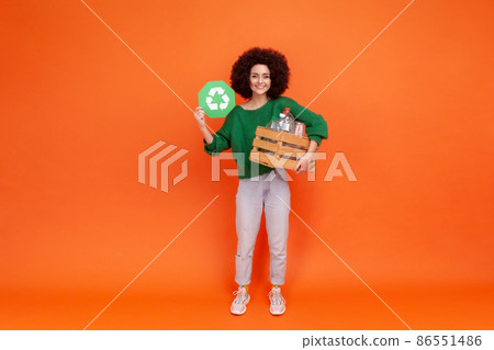 Full length portrait of woman with Afro hairstyle wearing green casual style sweater holding box with empty plastic bottler and showing green sign. Indoor studio shot isolated on orange background. Full length portrait of woman with Afro hairstyle wearing green casual style sweater holding box with empty plastic bottler and showing green sign. Indoor studio shot isolated on orange background. 86551486