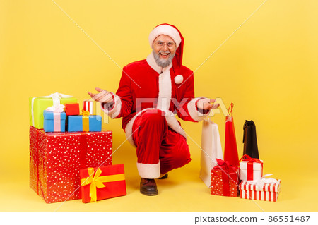 Portrait of elderly man with gray beard in santa claus costume showing many present boxes and shopping bags, being ready to congratulate with New year. Indoor studio shot isolated on yellow background 86551487