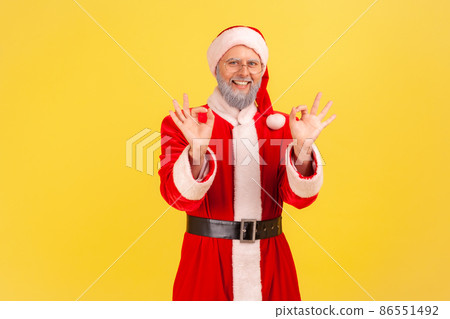 Portrait of satisfied elderly man with gray beard wearing santa claus costume showing okay sign with both hands, looking at camera toothy smile. Indoor studio shot isolated on yellow background. Portrait of satisfied elderly man with gray beard wearing santa claus costume showing okay sign with both hands, looking at camera toothy smile. Indoor studio shot isolated on yellow background. 86551492