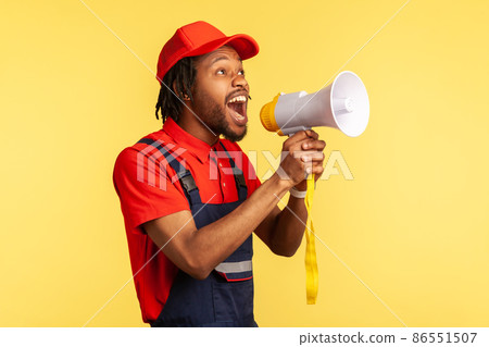 Portrait of bearded handyman wearing blue overalls and red cap screaming announcing about discounts of service industry or protesting. Indoor studio shot isolated on yellow background. 86551507