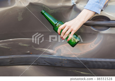 Unknown female hand holding bottle with beer white sitting behind wheel of broken car, road accident due to drunk driving, damaged side part of automobile after breakdown. Outdoor shot. 86551528