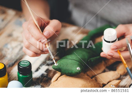 Handmade soft Doll frog. DIY gift crafting. Human hands hold frog soft toy. Needlework accessories on grey background. Handmade house decor. Process of making a vintage soft toy. Home workshop. 86551581