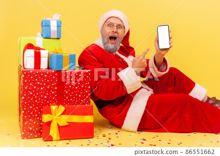 Astonished elderly man with gray beard wearing santa claus costume sitting pointing white blank screen of smart phone, surrounded with Christmas gifts. Indoor studio shot isolated on yellow background Astonished elderly man with gray beard wearing santa claus costume sitting pointing white blank screen of smart phone, surrounded with Christmas gifts. Indoor studio shot isolated on yellow background 86551662