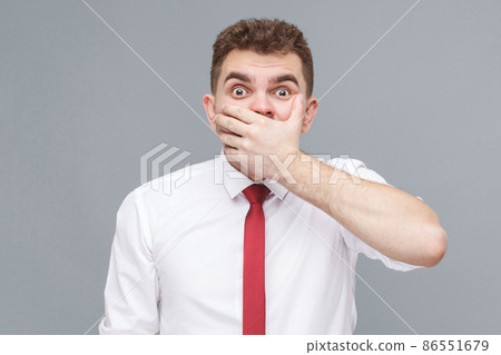 Portrait of young shocked man in white shirt and tie standing closing his mouth with hand and looking at camera with shocked face and big eyes. indoor isolated on gray background. Portrait of young shocked man in white shirt and tie standing closing his mouth with hand and looking at camera with shocked face and big eyes. indoor isolated on gray background. 86551679
