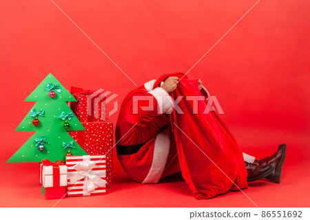 Unknown man wearing santa claus costume sitting on floor near decorative paper Christmas tree and gifts, putting head in bags with presents. Indoor studio shot isolated on red background. Unknown man wearing santa claus costume sitting on floor near decorative paper Christmas tree and gifts, putting head in bags with presents. Indoor studio shot isolated on red background. 86551692