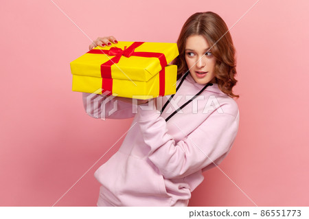 Portrait of curious teenager girl with brunette hair in hoodie secretly peeping inside wrapped yellow gift box, birthday present. Indoor studio shot isolated on pink background Portrait of curious teenager girl with brunette hair in hoodie secretly peeping inside wrapped yellow gift box, birthday present. Indoor studio shot isolated on pink background 86551773