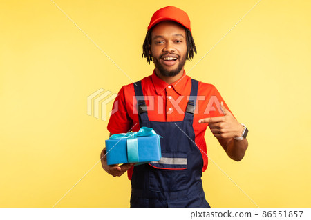 This present for you! Happy courier in overalls pointing at blue wrapped box, carrying birthday surprise, delivery service of purchases and gifts. Indoor studio shot isolated on yellow background. 86551857