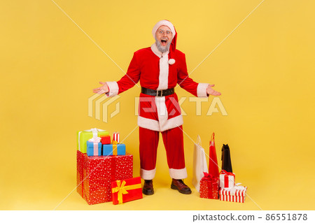 Full length portrait of elderly man with gray beard in santa claus costume standing with excited expression and raised arms near Christmas gifts. Indoor studio shot isolated on yellow background. Full length portrait of elderly man with gray beard in santa claus costume standing with excited expression and raised arms near Christmas gifts. Indoor studio shot isolated on yellow background. 86551878