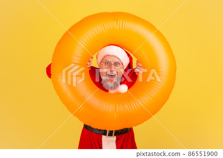Portrait of elderly man with gray beard wearing santa claus costume has amazed shocked expression, celebrating winter holidays in warm resort. Indoor studio shot isolated on yellow background. 86551900