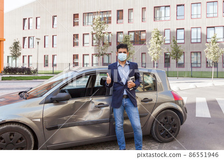 Portrait of happy brunette man wearing medical mask, jeans and jacket, holding tablet in hand and showing thumb up, insurance inspection crashed automobile after accident. Outdoor shot. 86551946