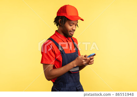 Side view portrait of positive deliveryman wearing uniform, using smartphone for taking new order for delivering, delivery service. Indoor studio shot isolated on yellow background. 86551948