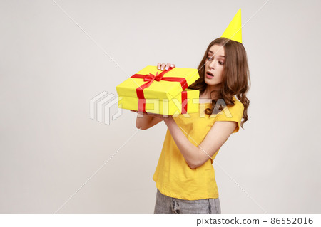 Portrait of curious amazed teenager girl in yellow T-shirt and party cone opening gift box, looking inside with surprised expression. Indoor studio shot isolated on gray background. 86552016
