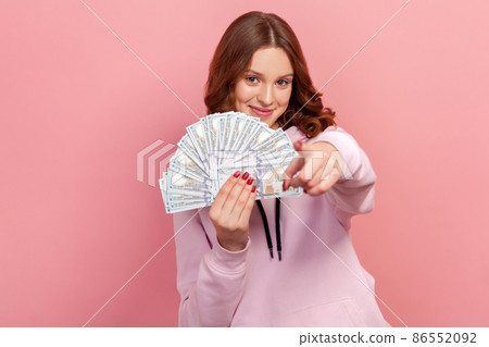 Portrait of joyful curly haired teenage girl holding bunch of dollar banknotes and pointing finger on camera, hey you gesture, winner. Indoor studio shot isolated on pink background Portrait of joyful curly haired teenage girl holding bunch of dollar banknotes and pointing finger on camera, hey you gesture, winner. Indoor studio shot isolated on pink background 86552092