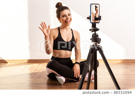 Trainer saying hello or bye, recording online tutorial of fitness exercises, using hone on tripod, wearing black sports top and tights. Full length studio shot illuminated by sunlight from window. Trainer saying hello or bye, recording online tutorial of fitness exercises, using hone on tripod, wearing black sports top and tights. Full length studio shot illuminated by sunlight from window. 86552094