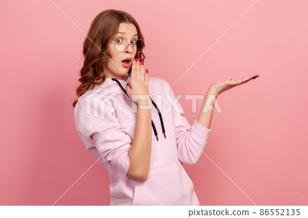 Portrait of shocked teen in eyeglasses closing mouth with hand and pointing right at copyspace, feeling surprised by unexpected information, omg gesture. Indoor studio shot isolated on pink background 86552135