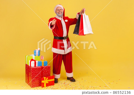 Positive elderly man with gray beard wearing santa claus costume holding shopping bags, pointing to camera, presents for Christmas holidays. Indoor studio shot isolated on yellow background. 86552136