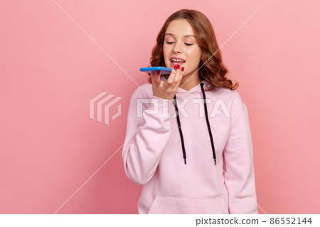 Portrait of brunette teen girl in hoodie talking to smartphone, using virtual assistant, ai intelligent technology, voice application, audio message. Indoor studio shot isolated on pink background 86552144