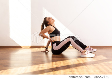 Side view of slim girl sitting touching sore spine after workout, has trauma, feeling discomfort, wearing black sports top and tights. Full length studio shot illuminated by sunlight from window. 86552180