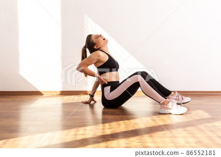 Side view of slim girl sitting touching sore spine after workout, has trauma, feeling discomfort, wearing black sports top and tights. Full length studio shot illuminated by sunlight from window. 86552181