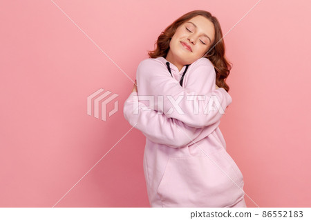 Portrait of selfish narcissistic brunette teen female in hoodie hugging herself and smiling with dreamy satisfied expression, love myself gesture. Indoor studio shot isolated on pink background 86552183