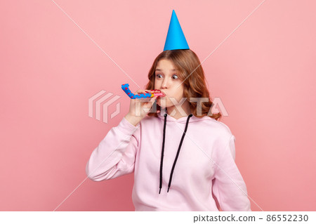 Portrait of funny curly haired teenage girl in hoodie with birthday hat blowing party pipe, celebrating holiday, fooling. Indoor studio shot isolated on pink background Portrait of funny curly haired teenage girl in hoodie with birthday hat blowing party pipe, celebrating holiday, fooling. Indoor studio shot isolated on pink background 86552230