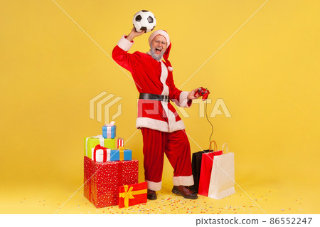 Full length of man with gray beard in santa claus costume raised arm with soccer ball, holding joystick, celebrating winning in football video game. Indoor studio shot isolated on yellow background. 86552247