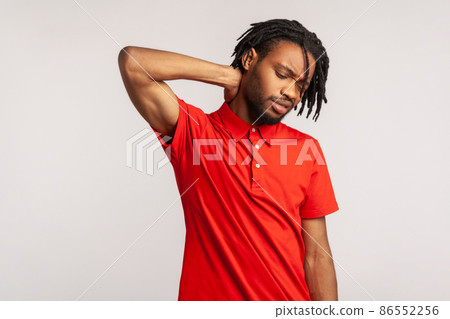 Sick bearded male wearing red casual style T-shirt, touching neck, feeling acute pain, suffering spine problems, osteochondrosis, frowning face. Indoor studio shot isolated on gray background. Sick bearded male wearing red casual style T-shirt, touching neck, feeling acute pain, suffering spine problems, osteochondrosis, frowning face. Indoor studio shot isolated on gray background. 86552256