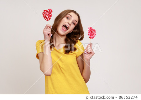 Portrait of cheerful playful woman in yellow casual T-shirt holding sweet round candy lollipop and looking at camera with excited childish expression. Indoor studio shot isolated on gray background. Portrait of cheerful playful woman in yellow casual T-shirt holding sweet round candy lollipop and looking at camera with excited childish expression. Indoor studio shot isolated on gray background. 86552274