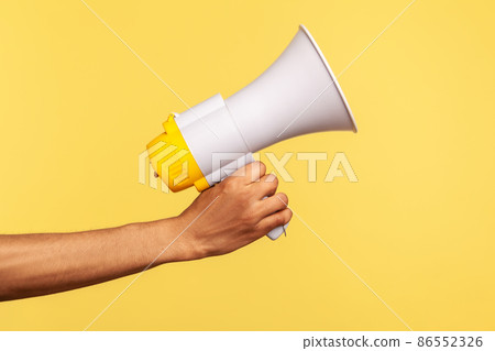 Profile side view closeup of human hand holding megaphone, announcing of advertisement. Indoor studio shot isolated on yellow background. 86552326