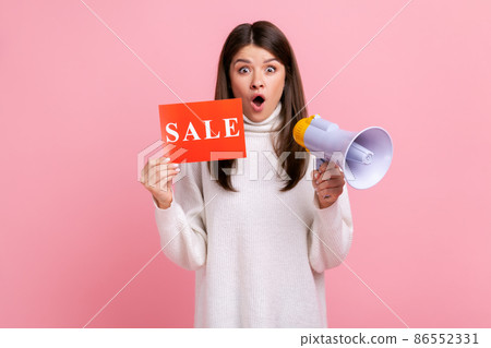 Shocked attractive female holding sale card and megaphone, announcing discounts in shopping mall, wearing white casual style sweater. Indoor studio shot isolated on pink background. 86552331