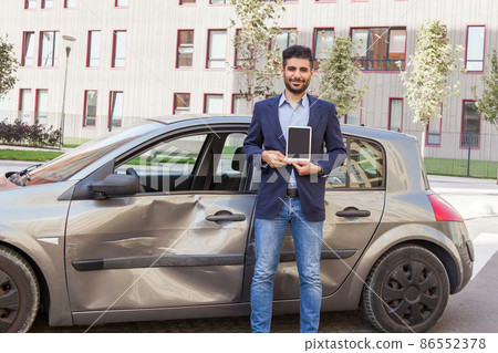 Young adult bearded man insurance inspector looking at camera with smile, holding showing tablet with empty screen for advertisement, standing near smashed automobile. Outdoor shot. 86552378