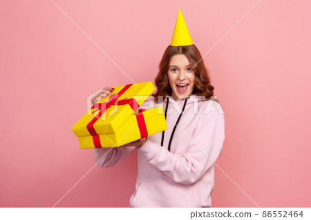 Portrait of surprised glad teenage girl in hoodie opening gift box and looking at camera with toothy smile, satisfied with present. Indoor studio shot isolated on pink background Portrait of surprised glad teenage girl in hoodie opening gift box and looking at camera with toothy smile, satisfied with present. Indoor studio shot isolated on pink background 86552464