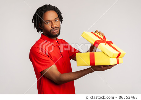 Disappointed man with dreadlocks in red casual T-shirt, holding unpacked gift box, has frustrated dissatisfied facial expression, upset with present. Indoor studio shot isolated on gray background. Disappointed man with dreadlocks in red casual T-shirt, holding unpacked gift box, has frustrated dissatisfied facial expression, upset with present. Indoor studio shot isolated on gray background. 86552465