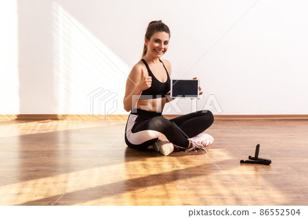 Sportive woman holds tablet with empty screen, showing thumb up, recommend online fitness course, wearing black sports top and tights. Full length studio shot illuminated by sunlight from window. 86552504