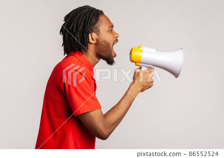 Profile of man with dreadlocks and beard wearing red casual style T-shirt, loudly screaming at megaphone, making protest, wants to be heard. Indoor studio shot isolated on gray background. Profile of man with dreadlocks and beard wearing red casual style T-shirt, loudly screaming at megaphone, making protest, wants to be heard. Indoor studio shot isolated on gray background. 86552524