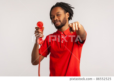Handsome guy man with dreadlocks wearing red T-shirt, pointing finger to camera, holding handset of red vintage landline phone, answering calls. Indoor studio shot isolated on gray background. 86552525