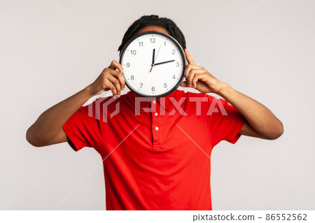 Unknown man with dreadlocks wearing red casual style T-shirt, hiding face behind big wall clock, time management, reminding of deadline. Indoor studio shot isolated on gray background. 86552562