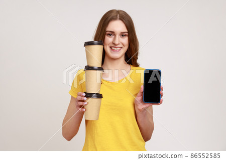 Delivery mobile app, internet advertise. Happy friendly teenager girl in yellow T-shirt holding stack of coffee cups and cellphone with mock up display. Indoor studio shot isolated on gray background. Delivery mobile app, internet advertise. Happy friendly teenager girl in yellow T-shirt holding stack of coffee cups and cellphone with mock up display. Indoor studio shot isolated on gray background. 86552585
