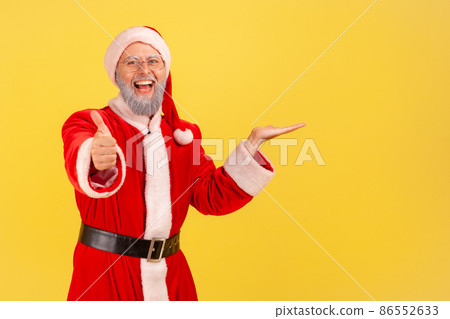 Glad happy elderly man with gray beard wearing santa claus costume presenting copy space for advertisement on his palm, showing thumb up, recommend. Indoor studio shot isolated on yellow background. Glad happy elderly man with gray beard wearing santa claus costume presenting copy space for advertisement on his palm, showing thumb up, recommend. Indoor studio shot isolated on yellow background. 86552633