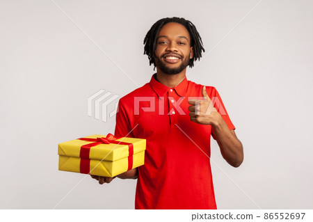 Bearded man with toothy smile wearing red casual style T-shirt, standing and holding yellow box and showing thumb up, satisfied with gift. Indoor studio shot isolated on gray background. 86552697