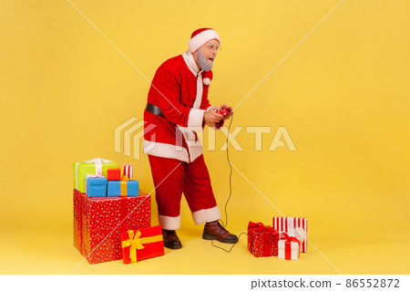 Full length portrait of elderly man with gray beard wearing santa claus costume playing video games in new year eve with concentrated expression. Indoor studio shot isolated on yellow background. Full length portrait of elderly man with gray beard wearing santa claus costume playing video games in new year eve with concentrated expression. Indoor studio shot isolated on yellow background. 86552872