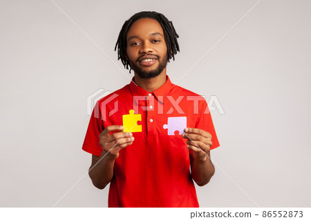 Calm smiling bearded man with dreadlocks wearing red casual style T-shirt, holding yellow and purple puzzle parts, solving problems and tasks. Indoor studio shot isolated on gray background. 86552873