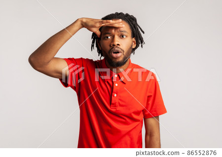 Astonished man with dreadlocks wearing red casual style T-shirt, holding hand over eyes and looking far away, watching horizon at long distance. Indoor studio shot isolated on gray background. 86552876