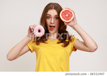 Playful excited teenager girl in yellow T-shirt holding half of ripe juicy grapefruit and round donut, looking camera with open mouth. Indoor studio shot isolated on gray background. 86552997