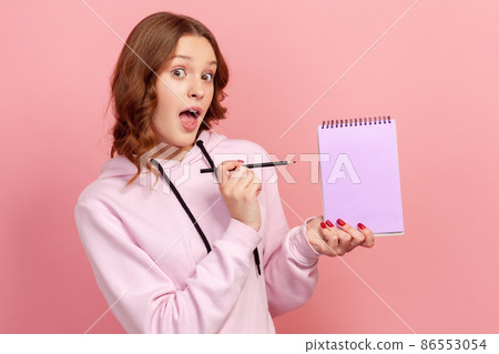 Portrait of surprised curly haired teenage girl in hoodie with opened mouth pointing pencil at empty notebook sheet, place for advertisement. Indoor studio shot isolated on pink background 86553054