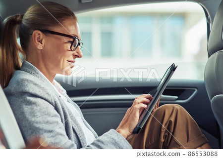 Side of a successful smiling business woman wearing eyeglasses using digital tablet, working while sitting on back seat in the car Side of a successful smiling business woman wearing eyeglasses using digital tablet, working while sitting on back seat in the car 86553088