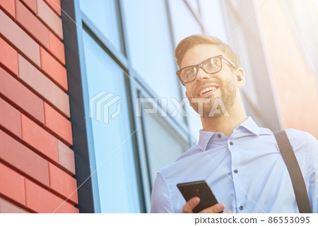 Young happy businessman listening to music with wireless earphones and mobile phone while walking outdoors 86553095