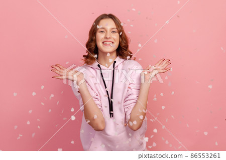 Cheerful young woman rejoicing falling heart shaped confetti, catching glitter with open palms and smiling at camera, girl celebrating valentines day. Indoor studio shot, isolated on pink background 86553261