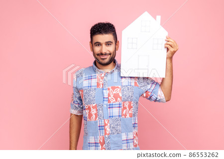 Attractive man with beard wearing blue casual style shirt holding paper house looking at camera with pleasant smile, dreaming of own home. Indoor studio shirt isolated on pink background. 86553262