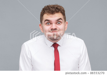 funny portrait of young man in white shirt and tie standing blowing his cheeks and looking at camera with funny face and big eyes. indoor isolated on gray background. 86553270